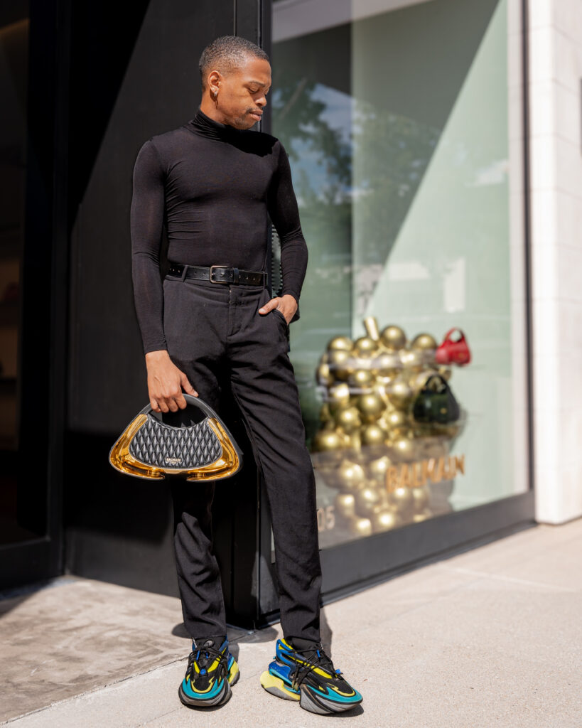 Minuit studio fashion photo - Street photo of a male Balmain model wearing a black Jolie Madame tote bag and multicolor Unicorn sneakers in neoprene and leather. picture tones are mainly gold and black